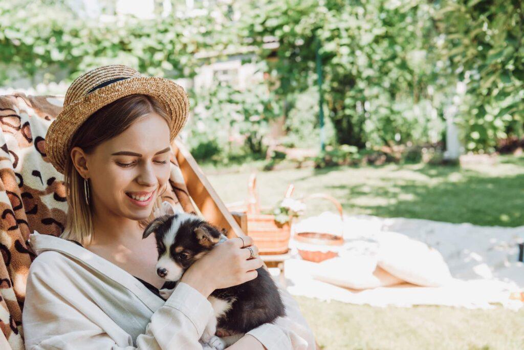 smiling blonde girl in straw hat holding puppy while sitting in deck chair in green garden 1